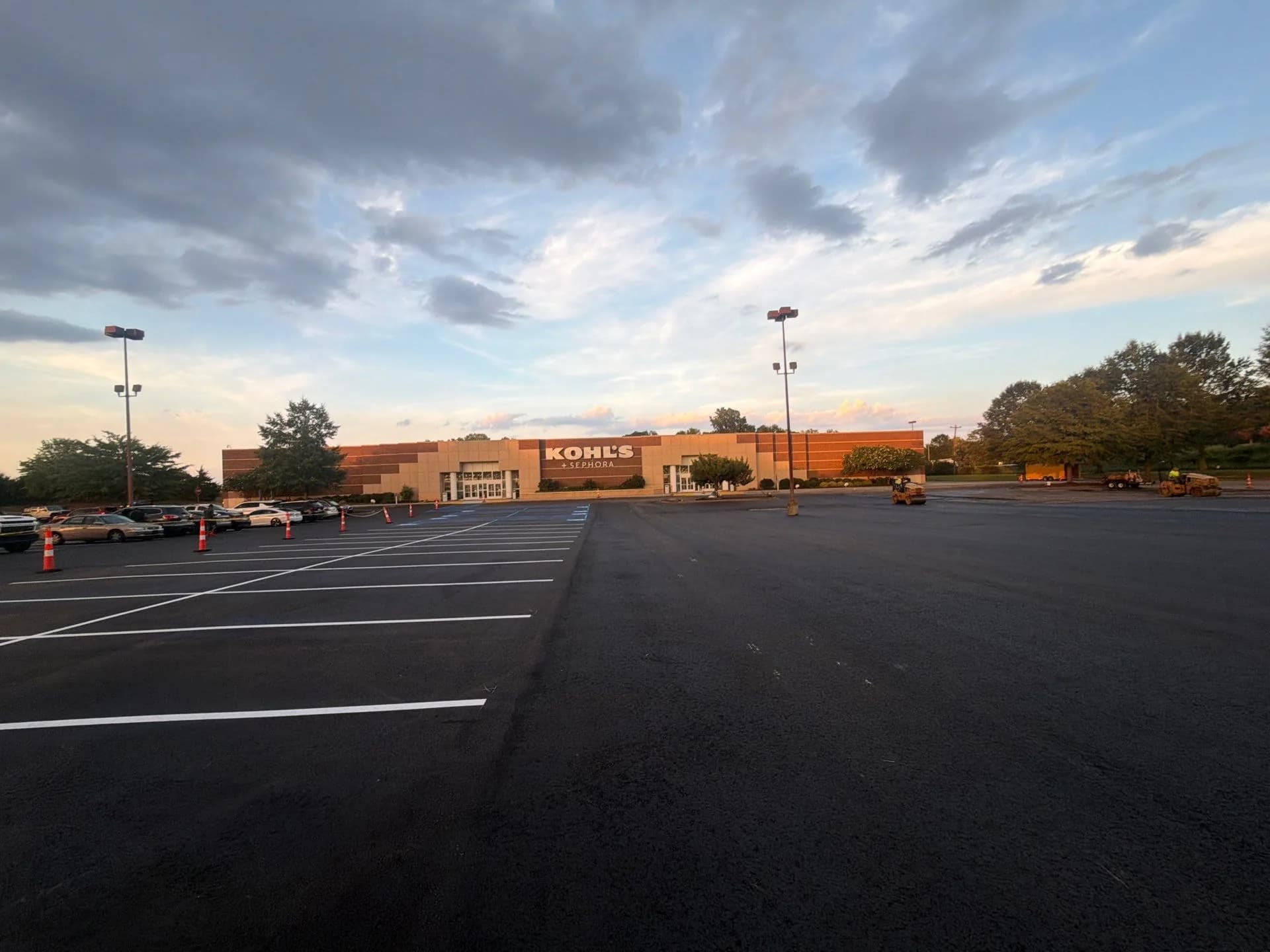 A big box retail parking lot at dusk with freshly paved asphalt and new white stall lines, cones separating the finished section from cars still parked near the store entrance