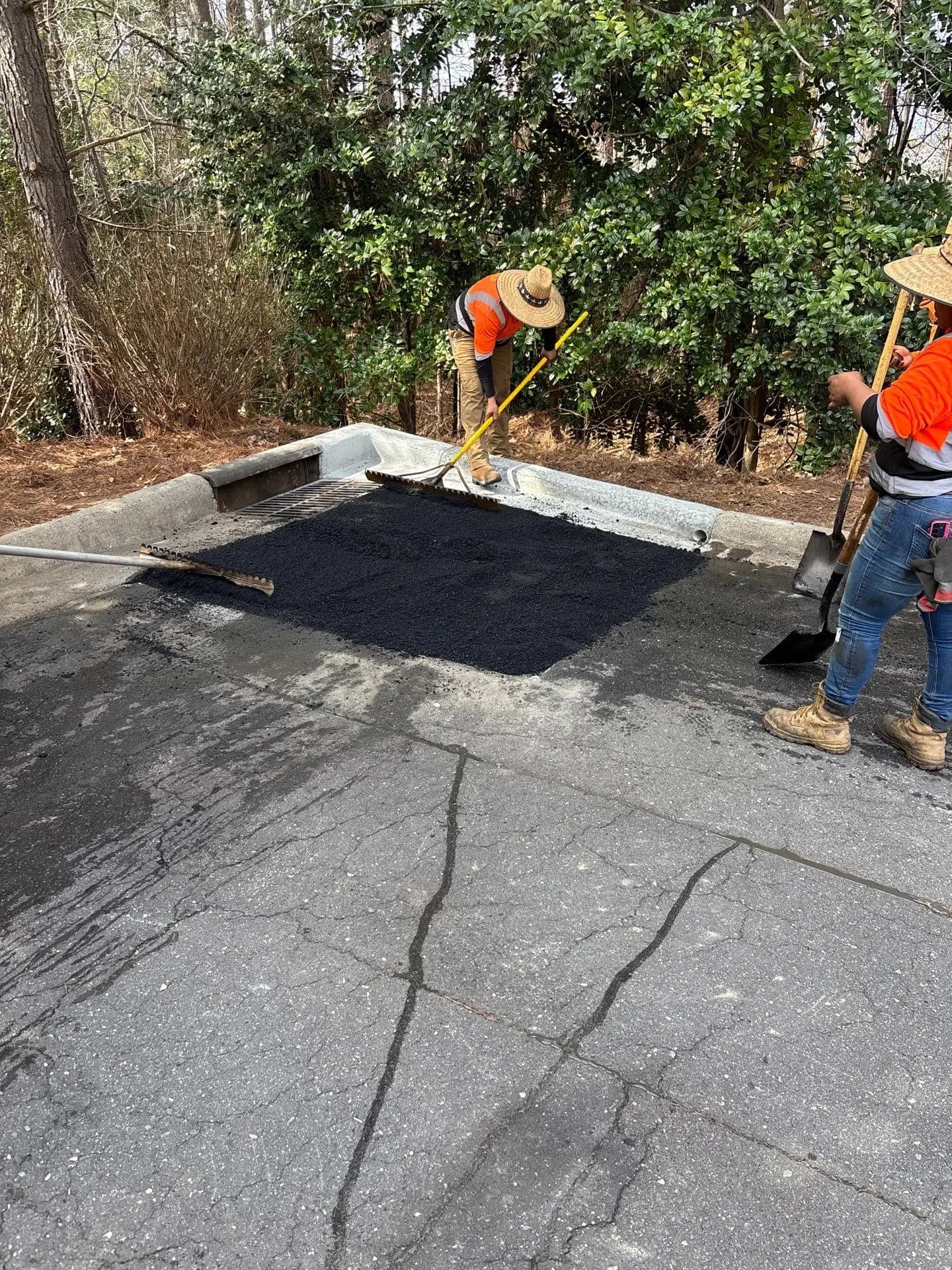 Crew raking hot mix into a cut-out patch beside a trench drain on a cracked lot