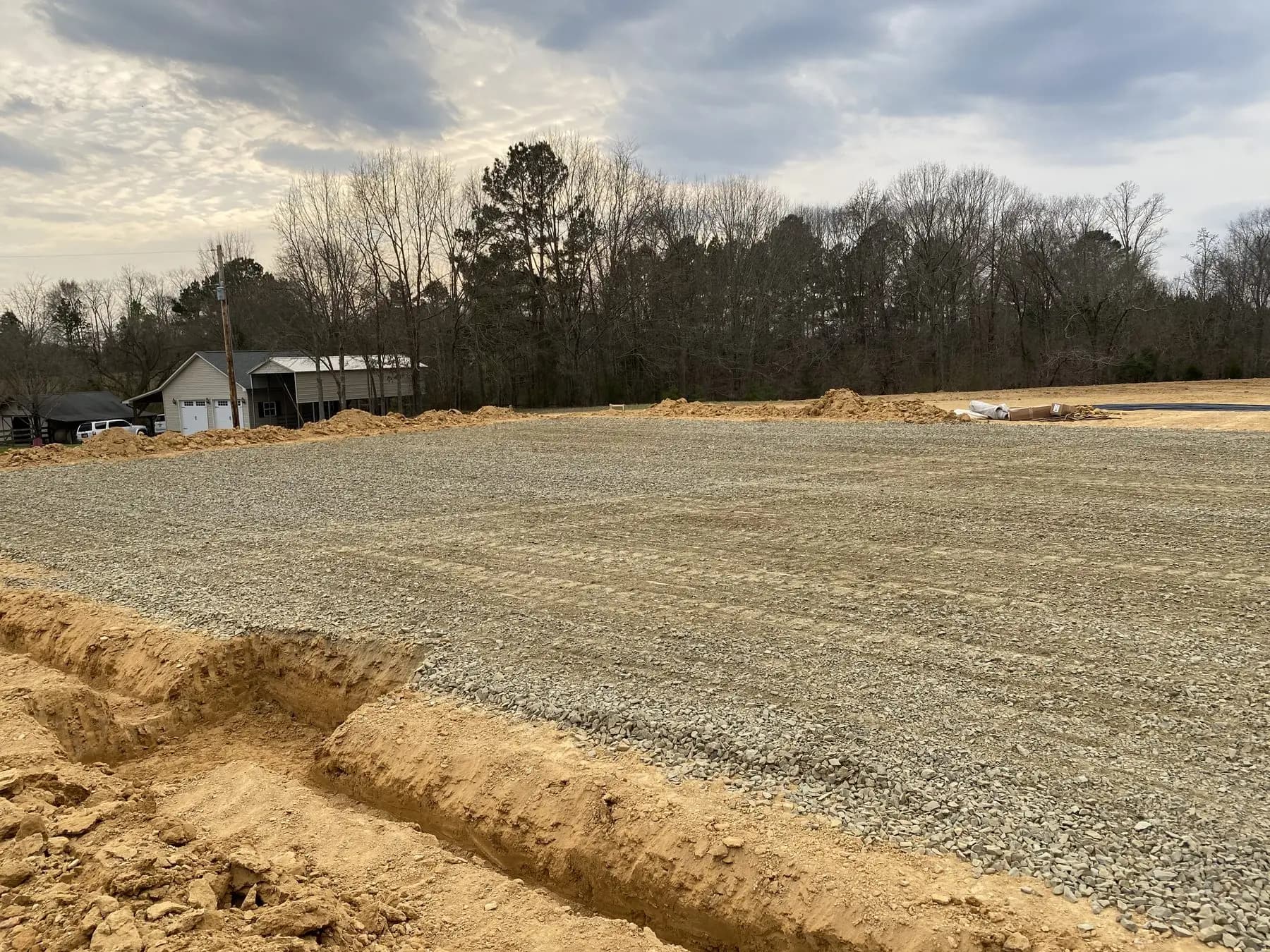 A graded and stone-based parking lot pad in Midland NC during the winter, with bare trees behind it and an excavated trench along the near edge, ready for asphalt
