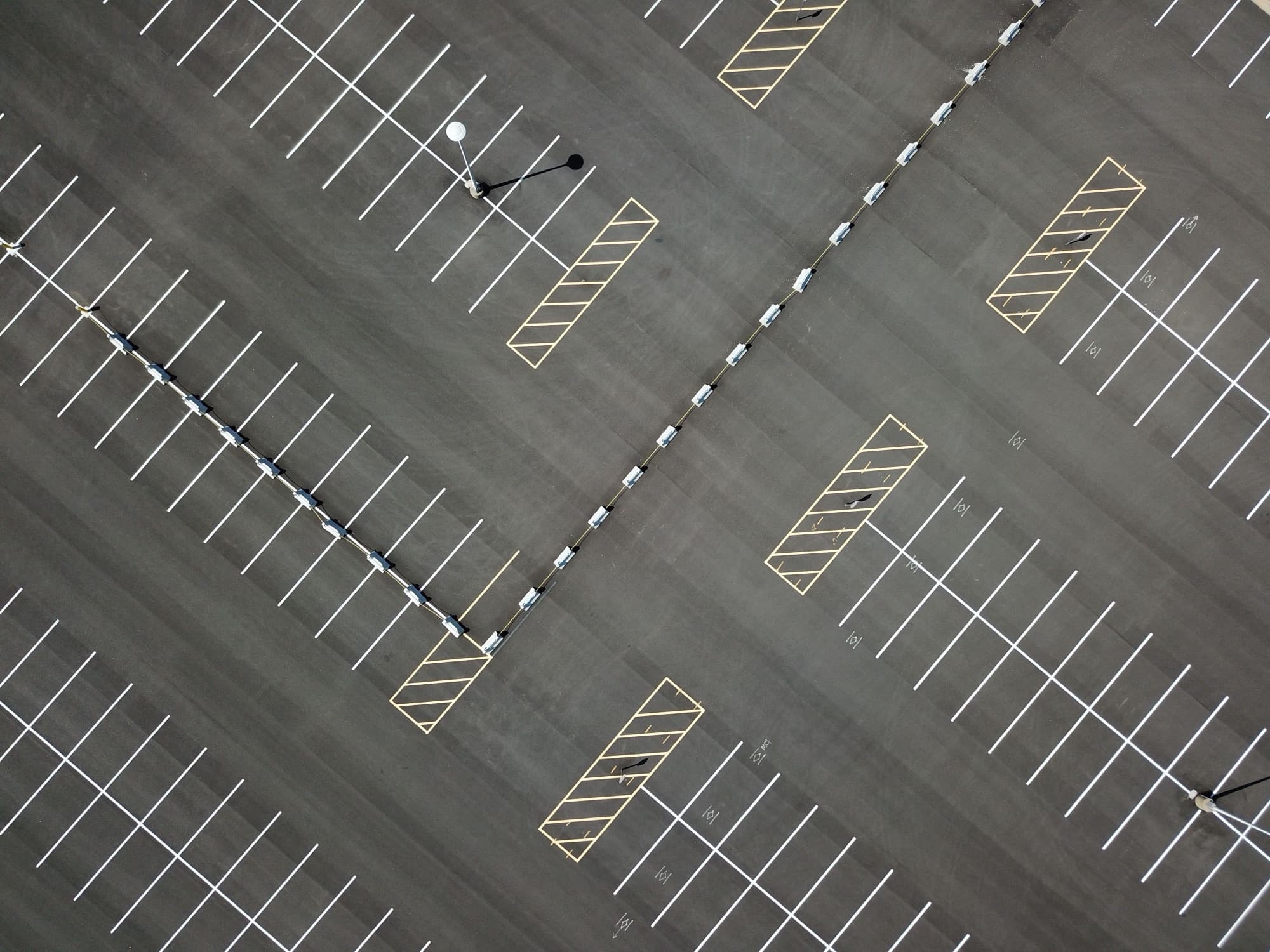 A large asphalt parking lot from directly above, crisply striped with hatched accessible bays